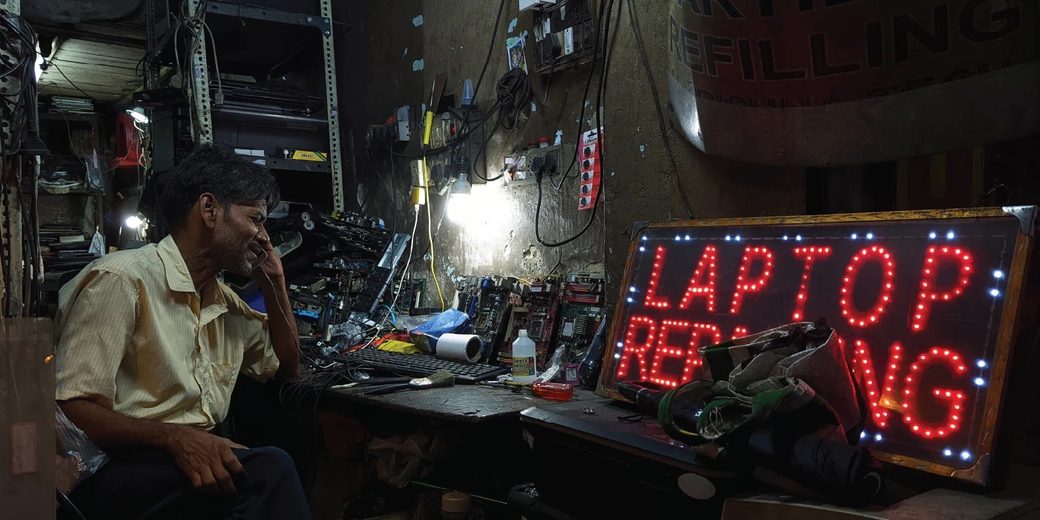 A Kolkata repair shop at night, a man on the phone at his workbench beside an LED sign spelling LAPTOP REPAIRING in red bulbs, a painted CARTRIDGE REFILLING board above