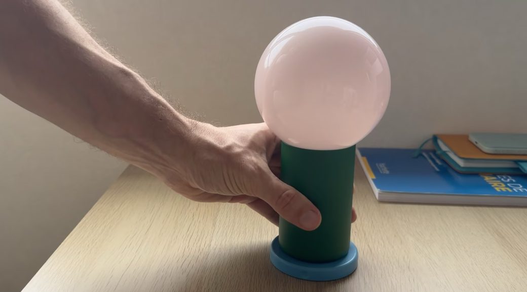 The Lightbulb Computer: a hand sets a large frosted bulb in a green battery base on a wooden desk, next to a stack of notebooks